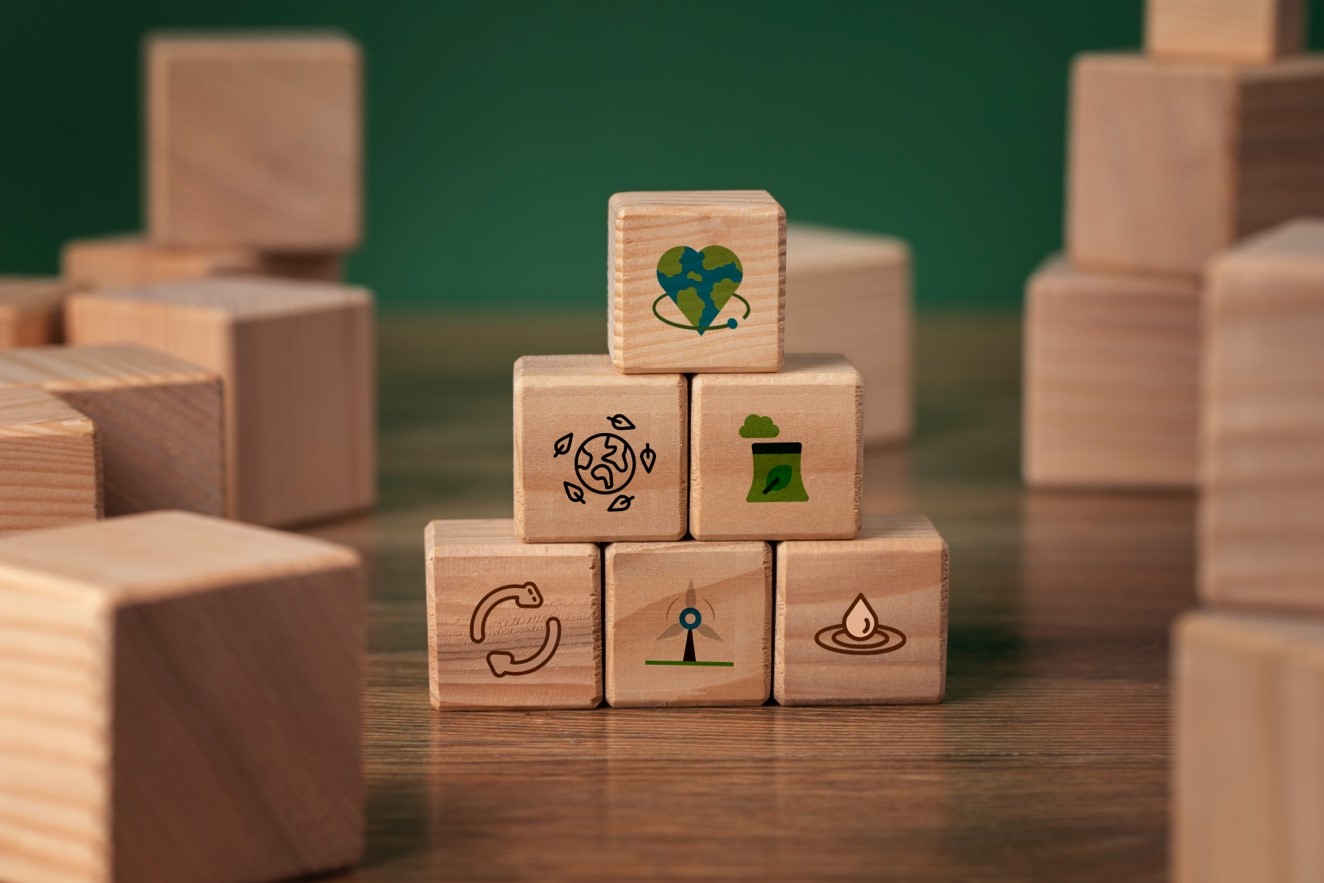 Wooden blocks stacked in a pyramid shape display eco-friendly icons, such as a globe, recycling symbol, and a leaf. The tone is clean and sustainable
