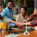 A diverse group of five colleagues smiling and stacking hands over a table with a small wind turbine model and plants, indicating teamwork and sustainability.