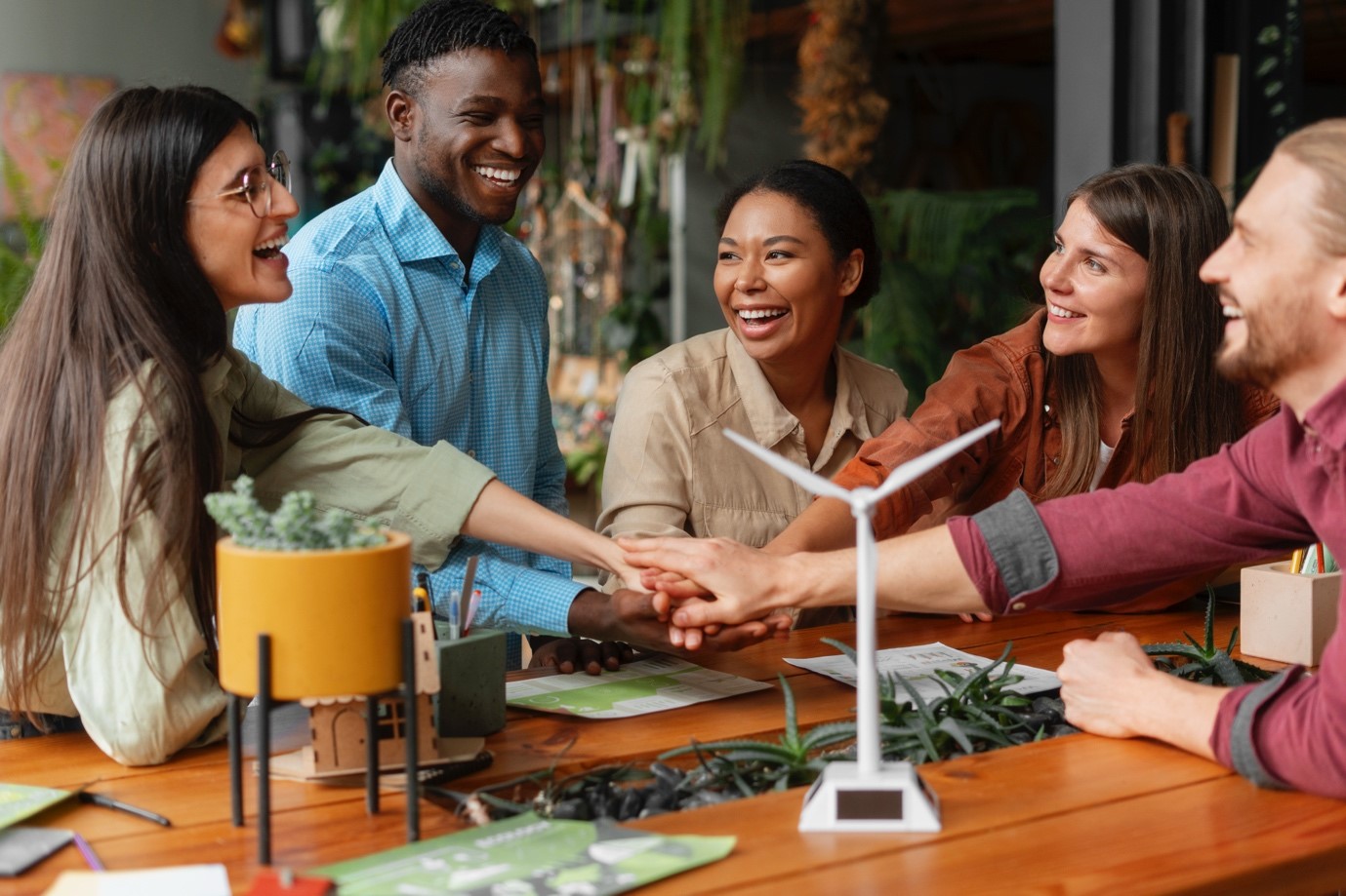 A diverse group of five colleagues smiling and stacking hands over a table with a small wind turbine model and plants, indicating teamwork and sustainability.